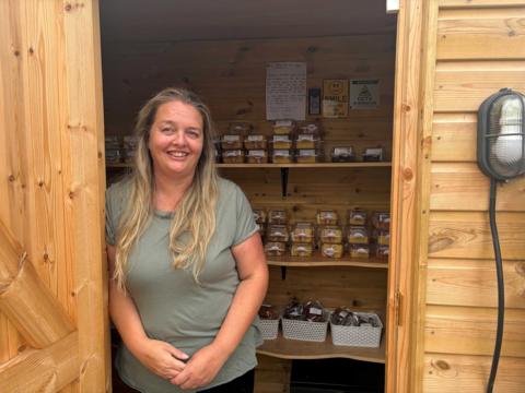 Emma Burton stands in the doorway of the Capel Cake Shed. Shelves behind her are filled with packaged cakes and traybakes.