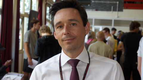 A man wearing a white shirt and a tie. He is also wearing a black lanyard and has short dark brown hair.
He is standing in a school and has a lot of people behind him 
