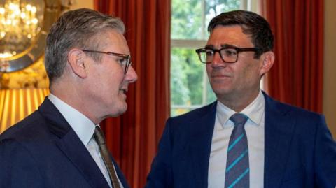 Keir Starmer talks to Andy Burnham as both wear suits and stand inside a room with curtains.