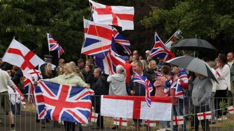 People hold flags as they gather near a hotel in Woolston as they protest over asylum seekers being housed in the hotel, in Warrington, on 30 August  30, 2025.