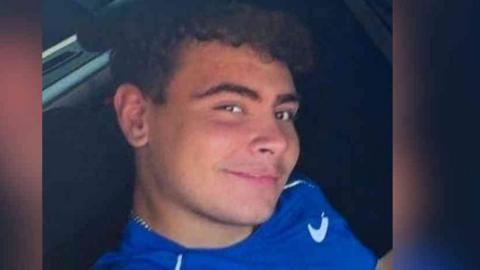 A teenager with curly brown hair and brown eyes smiles at the camera. He is wearing a blue top with a white trim and a branded tick.