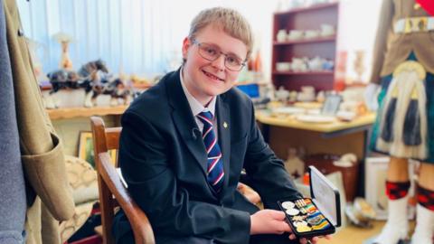 Charlie Elphinstone, wearing a blue blazer and striped tie, sits in his room holding a collection of medals