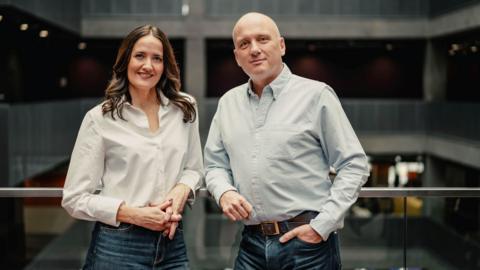 Laura Maciver and Martin Geissler casually lean on a glass railing inside the BBC Scotland building. They are both wearing jeans. Laura has long, dark hair and is wearing a white shirt. Martin has a bald head and is wearing a light blue shirt.