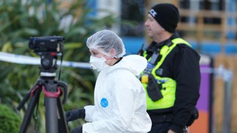 A person in a white forensics suit, hairnet and mask walking to the left. A man in a police uniform with a neon yellow vest is stood in the background. Some police tape and a camera on a tripod can also be seen.