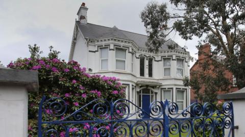 The former Kincora Boy's Home - a large, grey Victorian villa with bay windows on either side of its blue front door. The house is sited in a garden, behind a grey wall and decorative blue wrought-iron gates. There is a purple rhododendron plant in full blook in the garden. 
