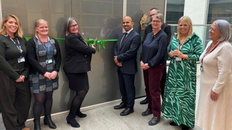 Smartly dressed people stood in a line, smiling at the camera. A woman in the middle is cutting a green ribbon over a grey door using a large pair of scissors.