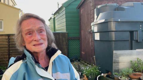 Pensioner Pauline Trubody stands in her garden next to a dark green heating oil tank, which is next to a brown shed. She has long grey hair and is wearing a blue and white coat.