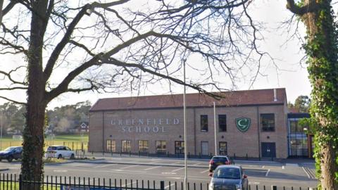 An external shot of Greenfield school which is a two storey brick building  with a red roof and the name of the school in large white letters on the walls. There is a car park in the foreground and playing fields are seen behind it.