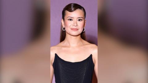 Katie Leung has long brown hair and wears a strapless black corset dress with silver hoop earrings