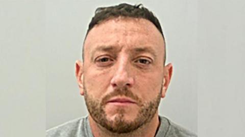 Police custody image of Jake Balmforth looking straight at the camera. He has short brown hair and stubble and is wearing a grey top.
