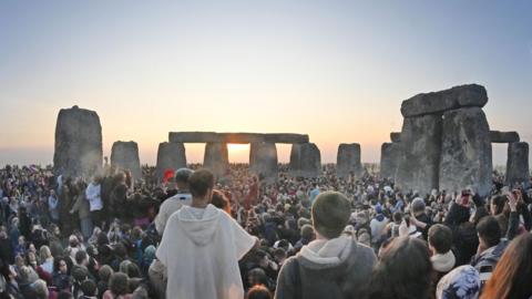 Big crowds around Stonehenge's famous big stones. One person is a wearing a white coat and stood slightly higher than the crowd
