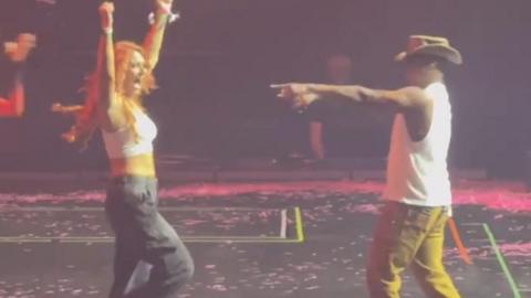 A woman with long red hair, a white tank top and black cargo trousers stands on a black stage stage with her arms in the air. To the right singer Ne-Yo, wearing a white vest top, mustard coloured trousers and a brown cowboy-style hat points towards her with his right hand. 