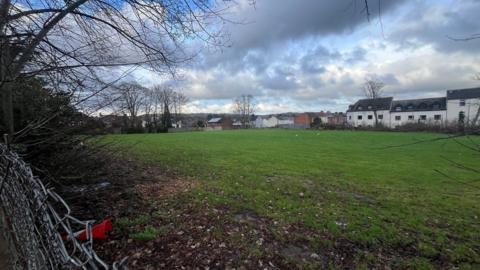 An area of grass surrounded by trees and hedgerows with houses visible in the distance