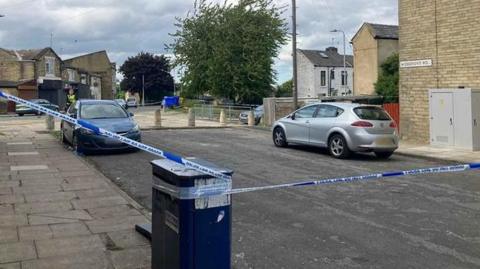 Police tape around a scene in a street. Two cars are pictured on the road, which comes to a dead end against a pavement. A police officer and houses can be seen in the distance.