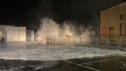 A large wave crashes over a metal barrier onto a waterfront walkway at night, lit by nearby buildings.