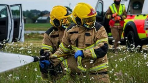 Two firefighters in brown overalls and yellow helmets kneeling in the grass and looking one way while squirting water the other, with two white vehicles in the background