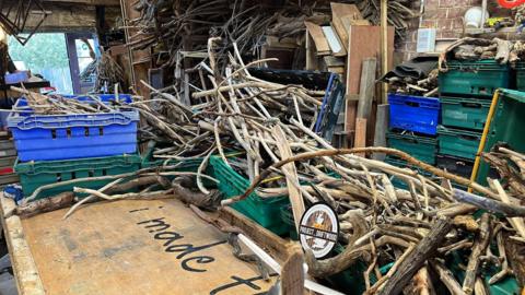 Studio full of driftwood piled up in boxes, with a sign reading "Project Driftwood" in the foreground.