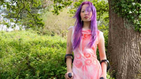 Josie Tang has purple hair. She is standing in woodland holding crutches, wearing a pink patterned dress. There is a large tree behind her, and green foliage.
