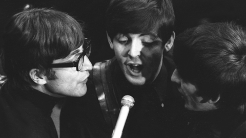 A black and white photograph of the Beatles recording for a ˿ radio programme. It is a close-cropped image of John Lennon, Paul McCartney and George Harrison singing around a microphone circa 1965