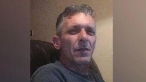 A man in his mid-50s with grey hair. He is sitting in an armchair and squinting at the camera.