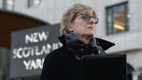 Dame Sally Davies holding a black folder outside New Scotland. Yard in London. She is wearing glasses, a black coat and a multi-coloured scarf.