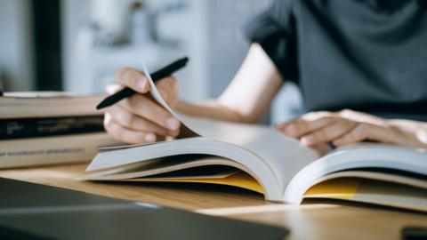 A young person, their face not visible, sits at a desk. They are holding a pen while turning a page of a book.