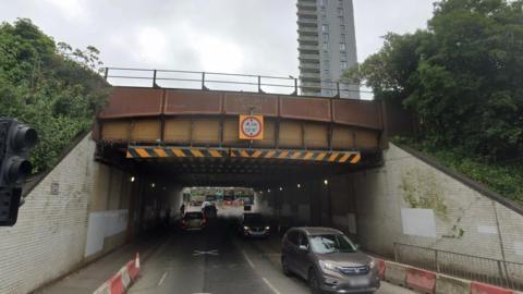 A road running under a brown bridge. The bridge is labelled Victoria Arch and has a height restriction sign attached.