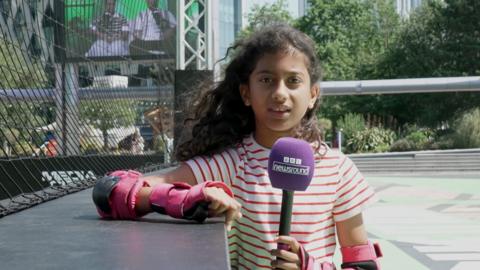 A young girl standing on a football pitch, holding a BBC Newsround microphone
