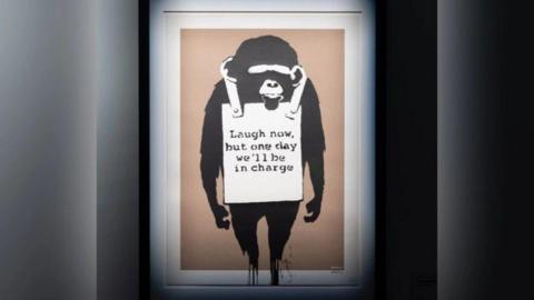 A Banksy artwork of a money with a white sign around its neck reading "laugh now, but one day we'll be in charge"
