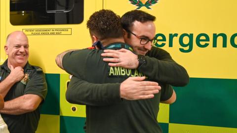 Patrick Mann hugs paramedic Matt Morgan while Jason Anderson looks on smiling 