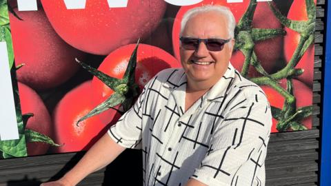 Tony Johnson is smiling at the camera and wearing a white shirt with black petterns on it.