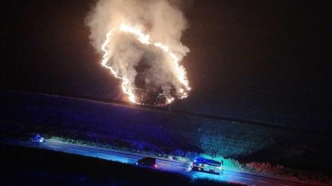 An aerial night-time image showing a concentrated area of burning grass producing flames and a large plume of smoke in a field near Stone Cross. A fire engine with blue lights is parked on a nearby road alongside other vehicles.