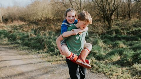 A young boy is carrying his young sister on his back. They are outside on path next to lots of tree and wild grass.