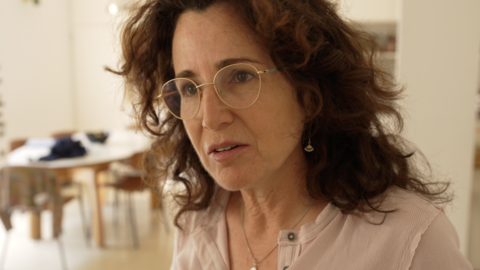 Liat Zvi is pictured from the shoulders up, in a domestic setting with a table in the background. She is a dark-haired woman with gold-rimmed glasses.