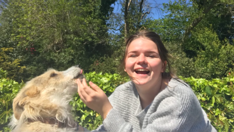 Sophie laughing outdoors whilst a blonde and white dog is licking her hand. 