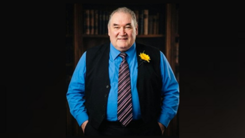 A man wearing a knitted, sleeveless cardigan over a blue shirt and a tie. He has a yellow flower attached to his cardigan. He is smiling while standing in front of a bookshelf. 