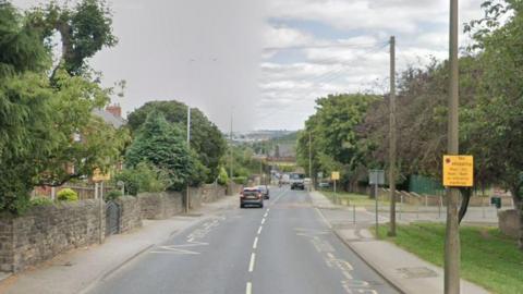 A road with walls and houses on the left hand side. On the right, trees and lampposts. A bridge in the distance.