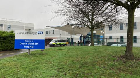 Welcome to Queen Elizabeth Hospital sign in front of white and grey building and tree without leaves