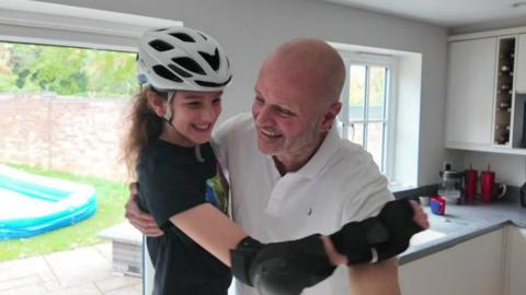 A father and his 11 year daughter hug each other in their kitchen, after the daughter gently rollerskates towards him. She has black curly hair, a black t shirt and wrist and elbow pads. He is bald and wearing a white polo shirt. Both are smiling