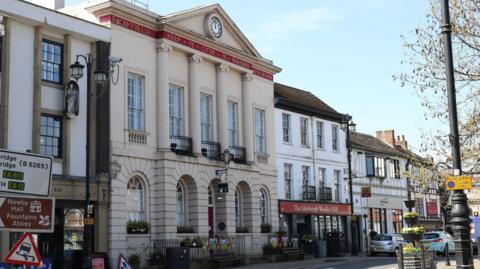 A historic town‑centre street with a neoclassical building featuring four columns and a clock in its pediment, alongside older shopfronts.