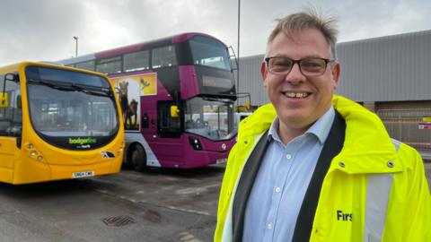 Doug Claringbold wearing black rimmed glasses, a blue shirt and a black blazer underneath a hi-vis yellow jacket. He is smiling at the camera, and standing in the Weston bus depot in front of two diesel buses which will soon be replaced with electric ones. 