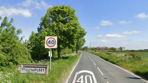 A road with a 40mph sign and the number 40 painted on the surface of the road. At the side of the road there is also a low brick block carrying the name of the village Stillingfleet written in black on a white background