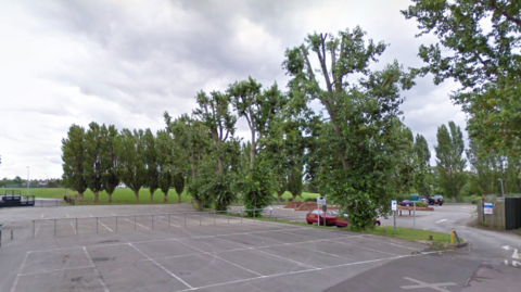 Google maps image of a car park behind a skatepark in Melksham, Wiltshire. Lots of empty car parking spaces can be seen, as well as trees. The field leading to the park can be seen in the distance. 