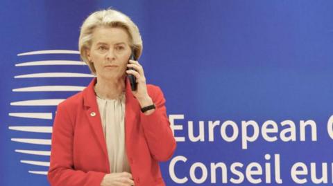 A woman in a pink jacket in front of a blue background holds a mobile phone to her ear