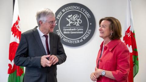 Mark Drakeford a Eluned Morgan yn siarad gyda'i gilydd gyda baneri Cymru yn y cefndir ac arwydd yn dweud 'Prif Weinidog Cymru'.