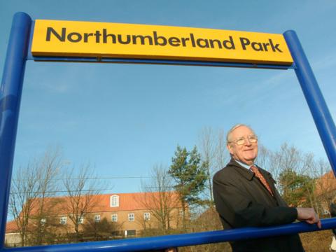 Tony Ridley standing under a sign for Northumberland Park Metro station in 2005. He has white hair and glasses. He is wearing a black rain coat, blue shirt and burgundy tie with blue and white dots. He is leaning on the frame of the sign and smiling. The frame is blue, with a yellow sign with the name of the station written in black.