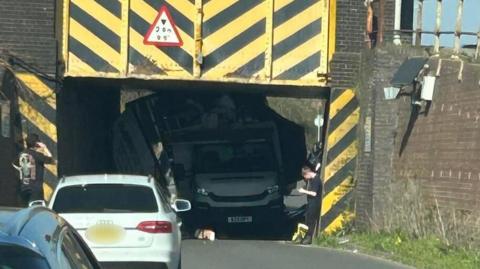 A white car parked in front of the entrance to a yellow and black striped railway bridge, inside which is a large removal van that has crashed into it.