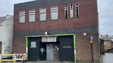 A building with brown bricks which is on two storeys and boarded up with some graffiti on it. 