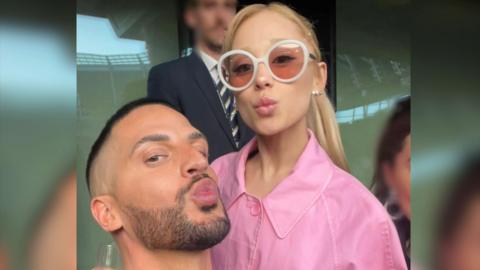 Carl Bembridge poses with the singer Ariana Grande. Carl and Ariana are both pouting and looking at the camera for a selfie. Ariana wears a pink jacket and white rimmed sunglasses.
