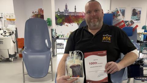 A man with a short greying beard sits in a hospital setting, holding a bag of donated blood and a certificate that reads: I have my 100th donation at Bradford Donor Centre. He wears a black polo short with a logo for the Kings Arms Pub on the chest.